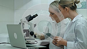 Researchers in labcoats are checking the samples on microscope in bright lab, using digital tablet and making notes on