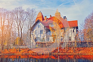 Residential building on river in autumn park. Beautiful vintage house in morning sunny sky. Yellow fall tree leaves on a