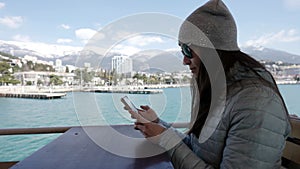 resort city near sea at winter day, adult woman is using smartphone sitting on open terrace of restaurant