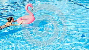 Resort wellness. Young sexy woman in bikini swimsuit with pink inflatable flamingo swimming in blue pool water. Straw