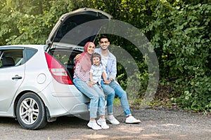 Rest and relax at road. Happy young arab male and female in hijab with small son sitting in car trunk