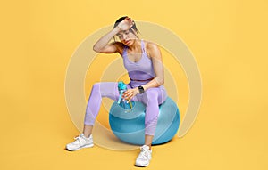 Rest and relax. Sporty tired black woman sitting on fitball with bottle of water after exercising, yellow background