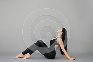 Rest between workouts. A pretty young woman with a good athletic form in a black jumpsuit is sitting on the floor on a gray backgr