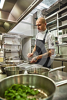 Restaurant kitchen. Vertical photo of concentrated chef in apron with several tattoos on his arms holding a frying pan