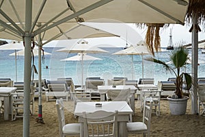 Restaurant tables and chairs setup under umbrella in white color and long chair in blue on Ornos sand beach with seaview