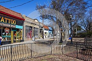 Restaurants and shops along a sidewalk with people, bare winter trees, lush green plants tall black light posts