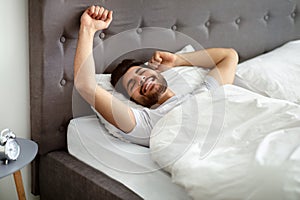 Rested arab man waking up and stretching hands, wellslept guy lying in bed, awakening in cozy bedroom and smiling