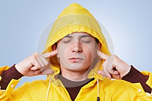 Restful attractive male closes eyes and plugs ears, tries to concentrate and not hear loud noise, wears yellow hood on head, isola