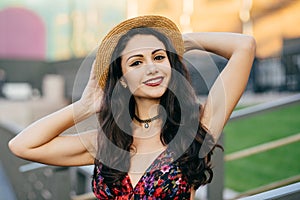 Restful brunette lady with beautiful make-up wearing summer hat and dress keeping her hand behind her head feeling pleasure while