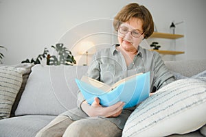 Restful reading. Portrait of thoughtful aged woman reading favorite literature at cozy home. She is lying on pillows on