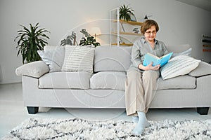 Restful reading. Portrait of thoughtful aged woman reading favorite literature at cozy home. She is lying on pillows on