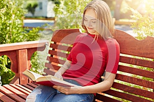 Restful woman with fair hair wearing red sweater and jeans sitting at wooden big bench holding book reading with pleasure. Student