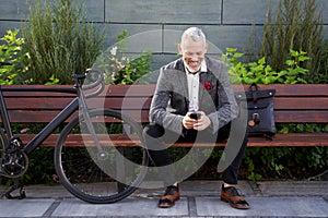 Resting. Successful middle aged business man in stylish suit texting, using mobile phone while sitting on the bench