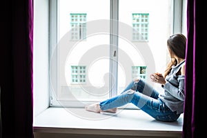 Resting and thinking woman. Calm girl with cup of tea or coffee sitting and drinking on the window-sill at home.