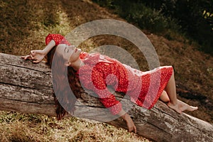 Resting young woman in red summer dress with dots lying on the fallen dry tree on the middle of wood