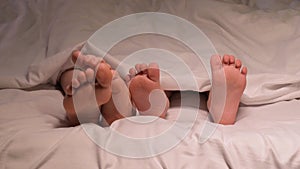 Restless legs of the boy son child and the mother female foot lie under the blanket on the sheet in the evening. Baby