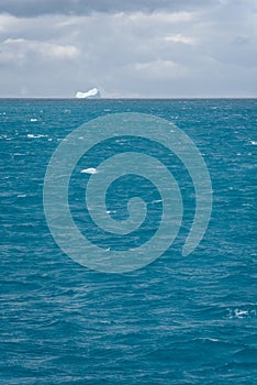 Restless nature background of choppy glacial melt water, floating ice chunks, stormy sky, and iceberg in background