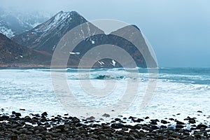 Restless winter blue sea with waves and snowy mountains background. Beach with stones in the foreground. Norway, Lofoten Islands