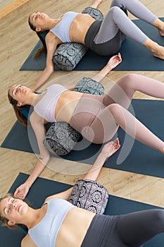 Restorative yoga with a bolster. Group of three young sporty attractive women in yoga studio, lying on bolster cushion