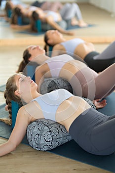 Restorative yoga with a bolster. Group of three young sporty attractive women in yoga studio, lying on bolster cushion