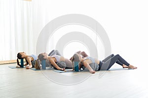 Restorative yoga with a bolster. Group of three young sporty attractive women in yoga studio, lying on bolster cushion