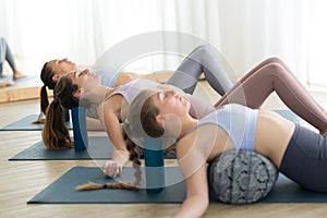 Restorative yoga with a bolster. Group of three young sporty attractive women in yoga studio, lying on bolster cushion