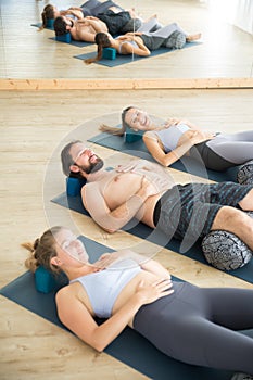 Restorative yoga with a bolster. Group of young sporty attractive people in yoga studio, lying on bolster cushion