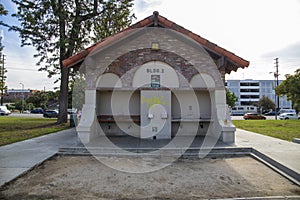 A restroom in the park with red brick and a sand pit in front with lush green trees and grass and cars parked along the street