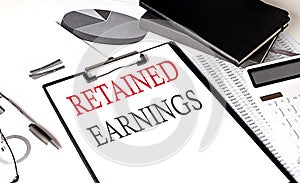 RETAINED EARNINGS text on paper clipboard with chart and notebook on withe background