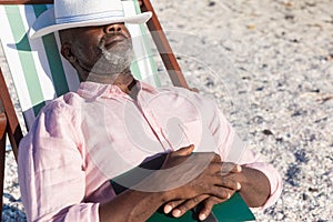 Retired african american senior man sleeping with book and hat over eyes on folding chair at beach