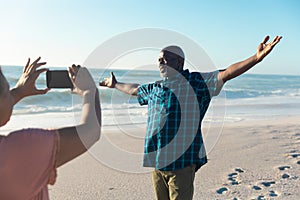 Retired african american senior woman photographing man with arms outstretched at beach