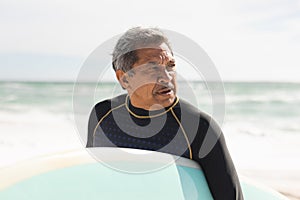 Retired biracial senior man looking away holding surfboard at beach against sky during sunny day