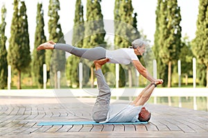 Retired couple of husband and wife performing advanced partner yoga pose in city park on sunny morning