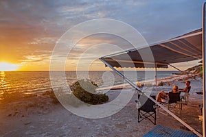 Retired couple sitting under the awning of a of caravan enjoying a wine next the beach with a golden sunset