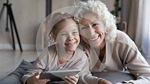 Retired grandmother and little granddaughter relax on pillows hold tablet