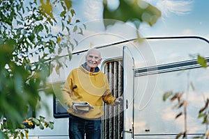 Retired man with book and newspaper in hands standing in door of his motor home