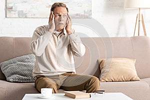 retired man with mental illness touching head while sitting on sofa.