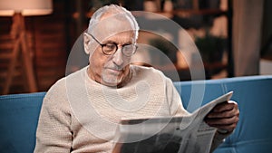 Retired man with newspaper sitting on couch in living room and reading, resting in evening in home