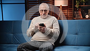 Retired man sitting on sofa in room and using mobile app on smartphone, portrait in home in evening
