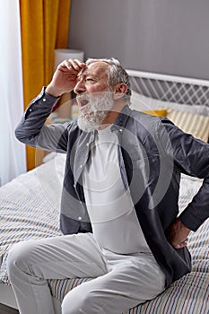 Retired man suffering from back pain at home, having difficulty in getting up from bed