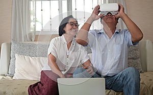 A retired man wearing a VR headset and his wife sitting side by side having a video call on the sofa in the house