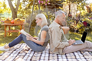 Retired married couple resting in garden, woman reading book while man using laptop, sitting back to back, side view