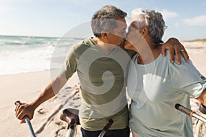 Retired multiracial senior couple kissing while standing with bicycles at beach during sunny day