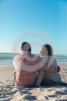 Retired senior african american couple talking while sitting at beach with copy space on blue sky