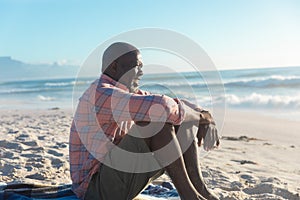 Retired senior african american man sitting on sand at beach looking away during sunny day