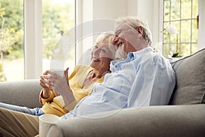 Retired Senior Couple Sitting On Sofa At Home Drinking Coffee And Watching TV Together