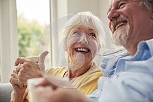 Retired Senior Couple Sitting On Sofa At Home Drinking Coffee And Watching TV Together