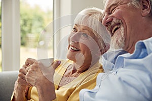 Retired Senior Couple Sitting On Sofa At Home Drinking Coffee And Watching TV Together