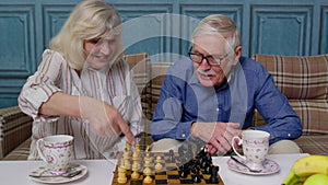 Retired senior couple talking drinking tea, playing chess in modern living home room lounge together