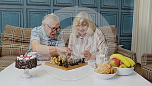 Retired senior couple talking drinking tea, playing chess in modern living home room lounge together